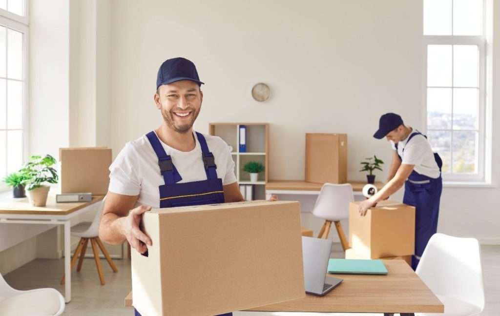 A smiling Smyrna mover holding a box in a customer’s home office while another tapes a box