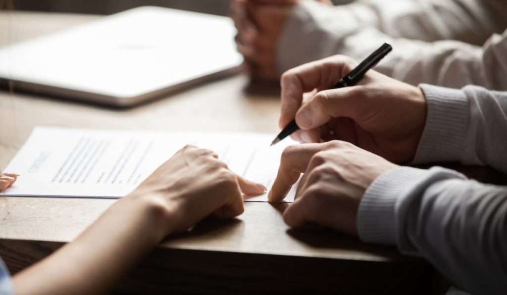 A person holding a pen to sign a lease, while another points at the document, and a third sits with hands folded 